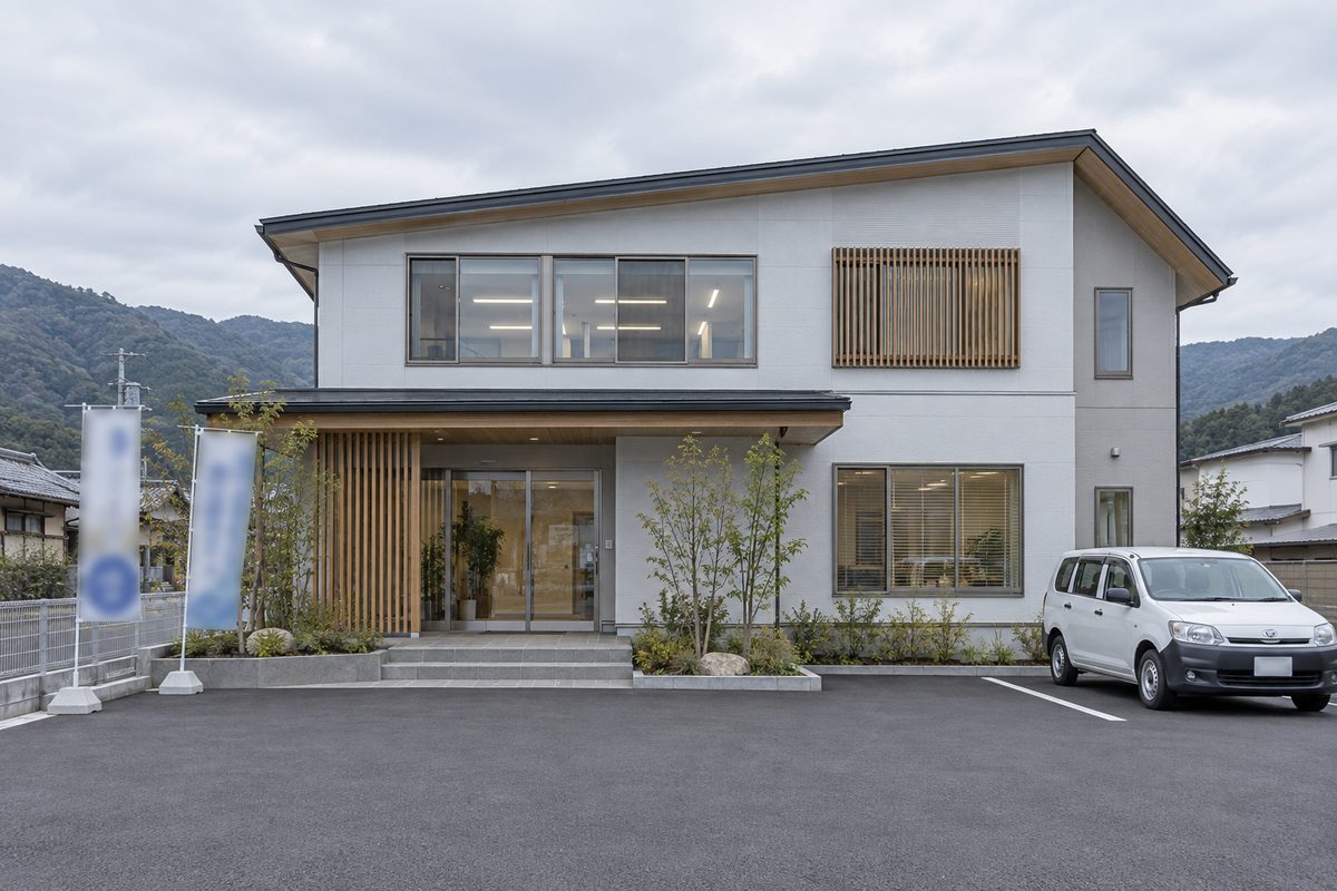 本社社屋の外観。2階建ての建物の前にのぼり旗が2本立ち、駐車場に社用車が1台停まっている。人物は写っていない。