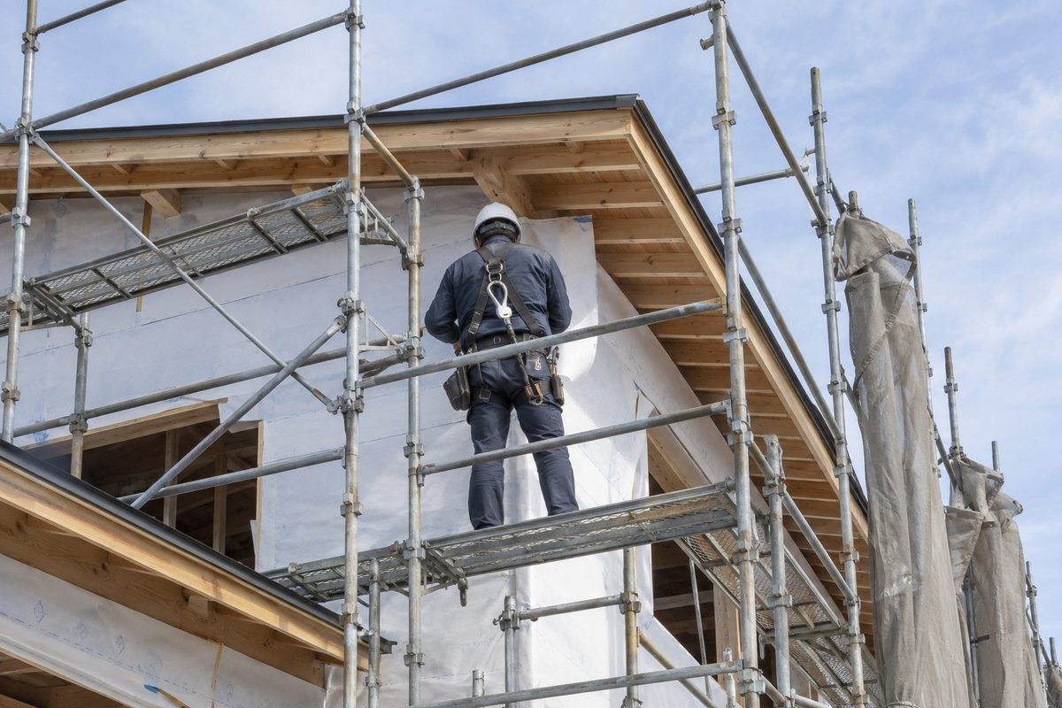 足場に立つ職人の後ろ姿。ヘルメットと安全帯を着け、建築中の外壁の前で作業している。顔は写っていない。